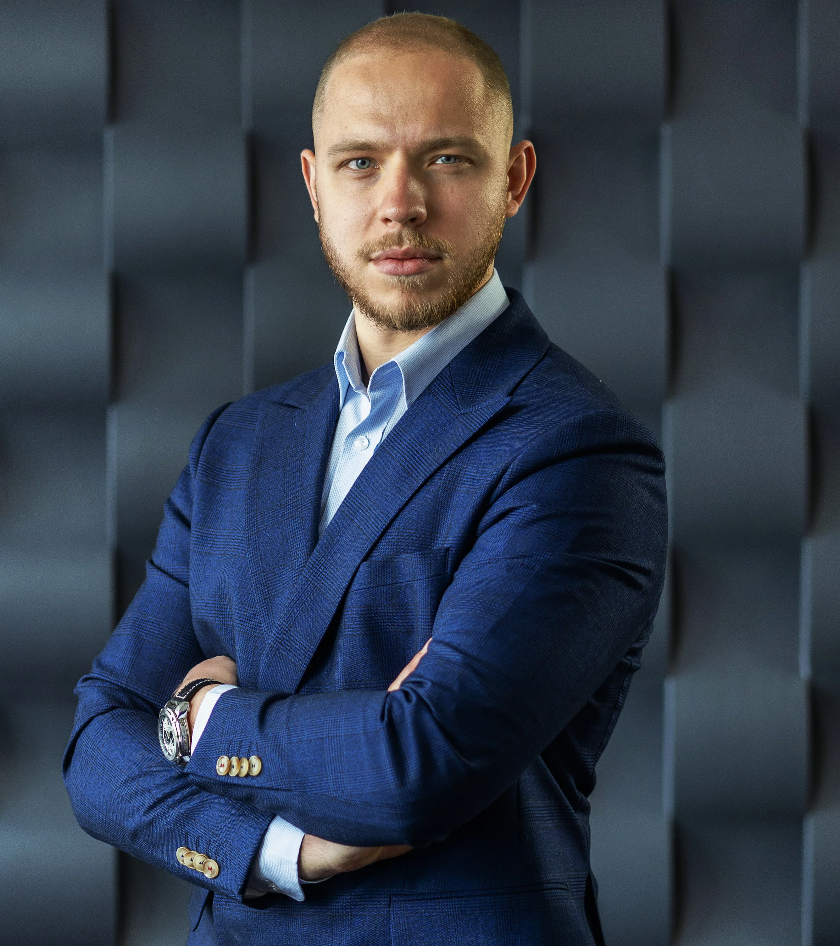 Tomasz Barnaś — portret biznesowy: mężczyzna w granatowym garniturze i jasnoniebieskiej koszuli, ze skrzyżowanymi rękami, na tle ciemnoszarej ściany akustycznej.
